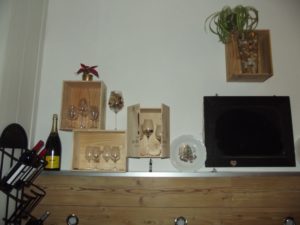 Die Weinkisten werden miteinander verschraubt und versetzt an die Wand montiert. Der Kelch in der Mitte ist mit Korken und einer Lichterkette gefüllt. Rechts an der Kiste befinden sich Türen, die aus einem Weinkisten-Deckel entstanden sind. Dafür müssen auf beiden Seiten zwei kleine Scharniere angebracht werden.